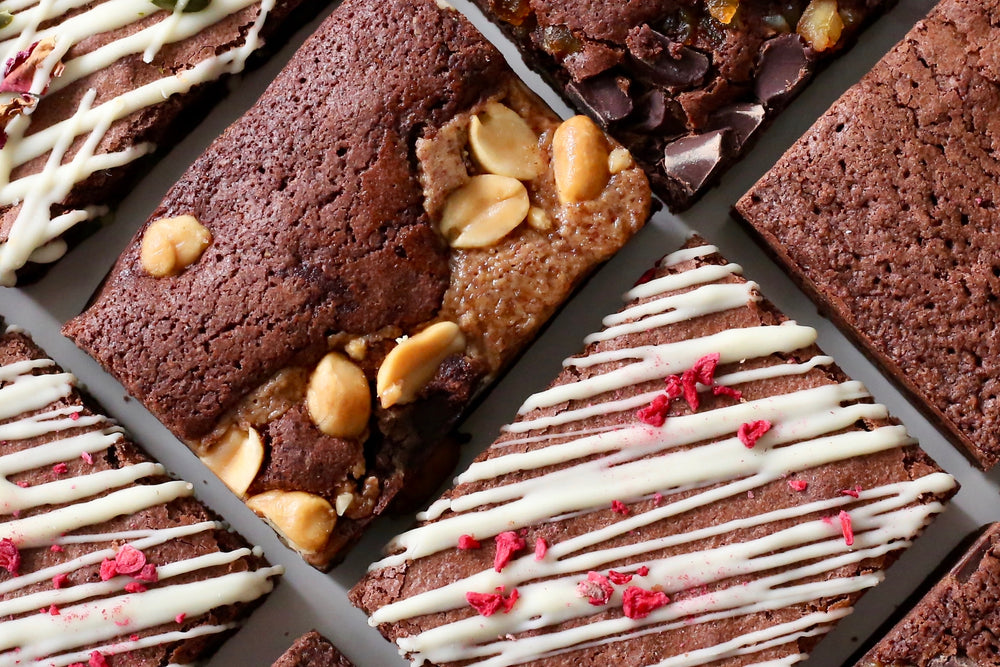 A close up picture of a selection of gourmet brownie slices.  Shows a brownie decorated with peanut butter and peanuts, a brownie with white chocolate drizzled and raspberry crumb on top.  An orange chocolate chip brownie in the corner.